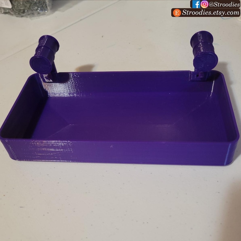 May include: A purple plastic tray with two small, round, cylindrical handles on either side. The tray is empty and appears to be made for holding small items.
