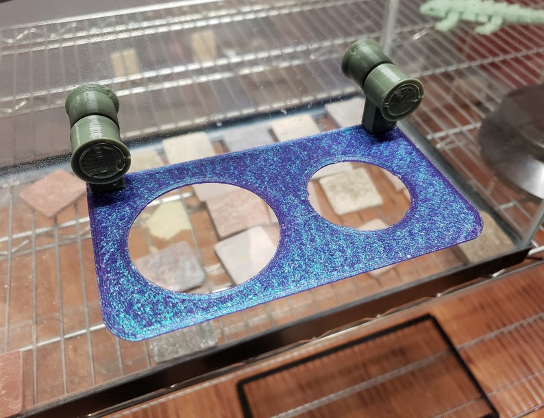 May include: A blue and green 3D printed reptile feeder with two circular openings. The feeder has two green clips on the top for attaching to a terrarium.