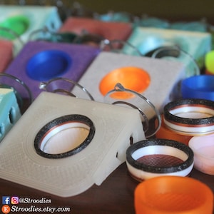 May include: A collection of 3D printed speaker stands in various colours, including purple, blue, orange, and white. The stands feature a circular opening for a speaker and a raised platform for stability. The stands are made of a smooth, textured plastic.