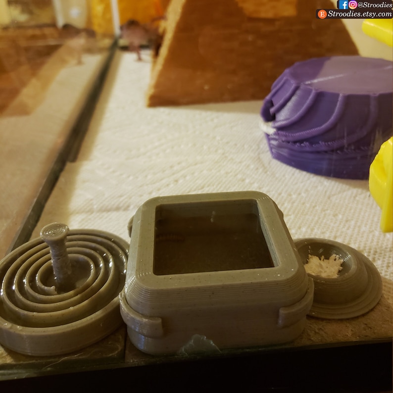 K&ouml;nnte beinhalten: Drei 3D-gedruckte Reptilien-Gehege, eines ist eine quadratische Box mit Deckel, eines ist eine runde Schale mit einem Mittelpfosten und das andere ist eine runde Schale mit einem erh&ouml;hten Rand.