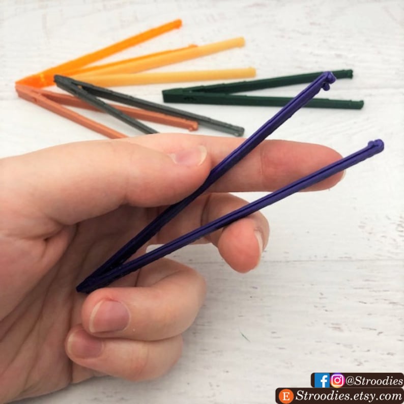May include: A set of colorful plastic tweezers, with one pair of purple tweezers held in a hand. The tweezers are designed for crafts and small tasks.