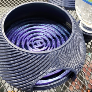 May include: A dark blue, spherical container with a textured, ribbed exterior. Inside, a swirling, spiral design in shades of purple and blue is visible. A clear glass is partially visible on the right.