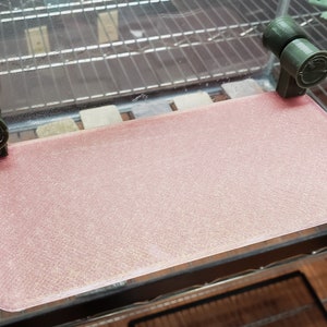May include: A pink, textured, rectangular glass shelf with two green plastic brackets on either side.