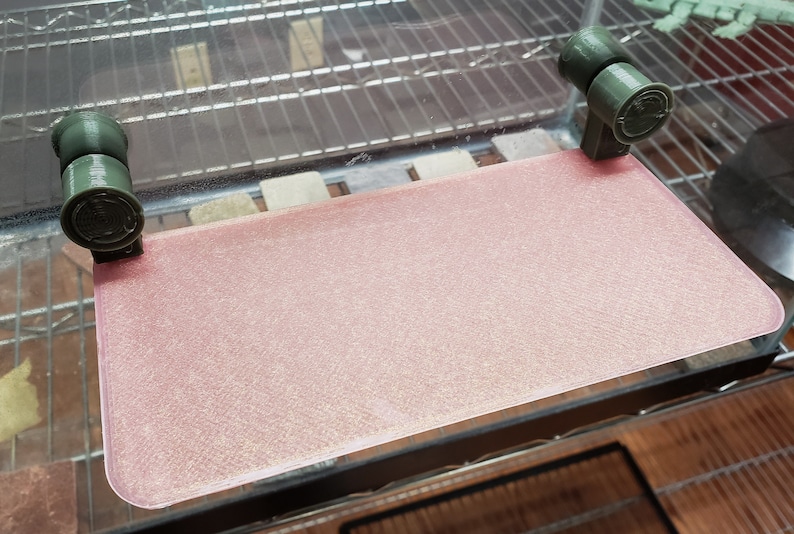 May include: A pink, textured, rectangular glass shelf with two green plastic brackets on either side.