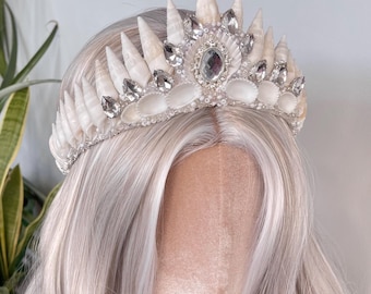 May include: A white seashell crown with rhinestones and pearls. The crown is on a white wig.
