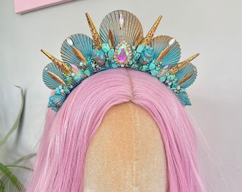 May include: A turquoise and gold mermaid crown with seashells, pearls, and rhinestones. The crown is on a pink wig mannequin.