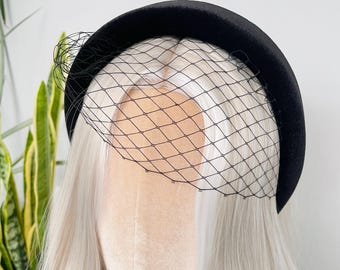 May include: A black velvet headband with a delicate black netting veil. The headband is thick and rounded, and the veil is attached to the front. The headpiece is displayed on a mannequin head with long, wavy, silver hair.