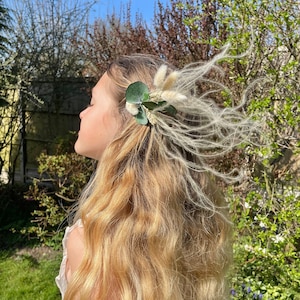 May include: A young girl with long blonde hair wearing a white dress and a floral hair clip with dried grasses and green leaves.