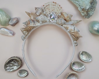 May include: A white headband decorated with seashells and pearls. The headband has a large iridescent jewel in the center.