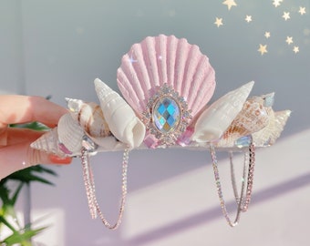 May include: A pink seashell crown with a large iridescent gem in the center, surrounded by smaller seashells and two dangling rhinestone chains.