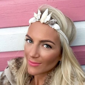 May include: A woman with long blonde hair is wearing a white seashell headband. The headband is made of a variety of seashells, including some that are white and some that are brown. The woman is wearing a brown and white fur jacket.