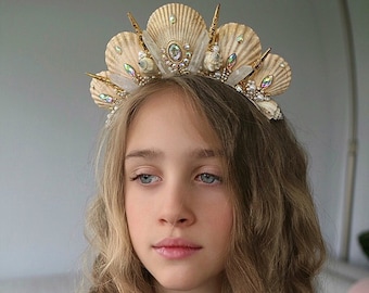 May include: A seashell crown with gold accents and iridescent gems. The crown is made of cream-colored seashells and is adorned with small pearls and gold-colored spikes. The crown is worn on a person's head.