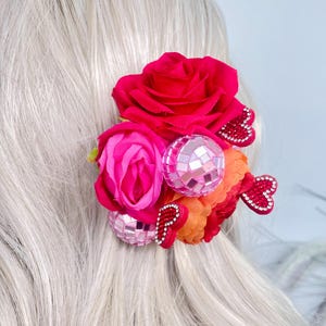 May include: A vibrant hair accessory featuring a cluster of red and pink fabric roses, a disco ball, and orange fabric accents. Two red heart-shaped embellishments with rhinestones add detail. The accessory is set against a backdrop of light-colored hair.