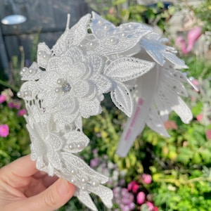 May include: White floral headband with pearl accents. The headband is made of delicate lace and features a large white flower with a rhinestone centre.