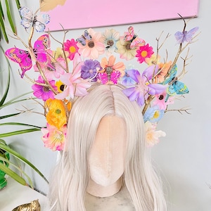 May include: A floral headpiece with a wig base, featuring colorful flowers and butterflies. The headpiece has gold-painted branches and a variety of flowers in shades of pink, yellow, purple, and blue. Butterflies in various colors are attached to the branches.