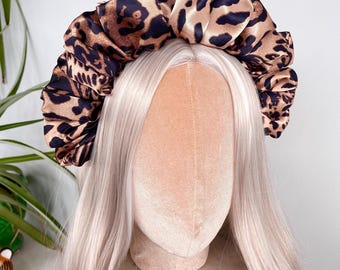Puede incluir: Una diadema de satén con estampado de leopardo. La diadema es marrón con manchas negras y se muestra en una cabeza de maniquí con cabello largo y claro. El fondo es una pared rosa claro y una superficie de mármol.