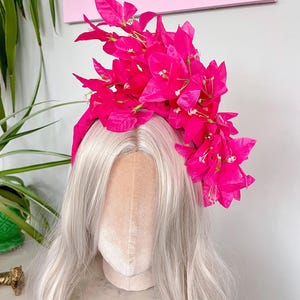 May include: A vibrant pink floral headband adorns a mannequin head with long, silver-gray hair. The headband features bright pink bougainvillea flowers, creating a striking contrast against the hair. The background includes a pink canvas and greenery.