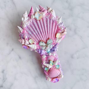 May include: A decorative hand mirror featuring a seashell design. The mirror is adorned with various seashells, pearls, and iridescent gems in shades of pink, purple, and white. A butterfly charm is centered on the mirror.