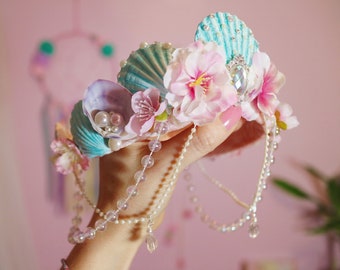 May include: A mermaid crown made of seashells, pearls, and pink flowers. The crown is decorated with iridescent beads and a crystal-like jewel.