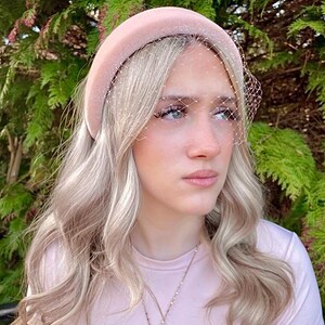May include: A light pink velvet headband with a matching veil. The headband is worn by a person with blonde hair and a pink shirt. A gold cross pendant necklace is also visible.