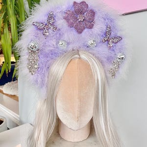 May include: A lavender halo crown featuring soft feathers, a central glittery purple flower, two sparkling butterfly embellishments, and dangling silver disco ball accents with clear crystal clusters attached to the base of the headpiece.