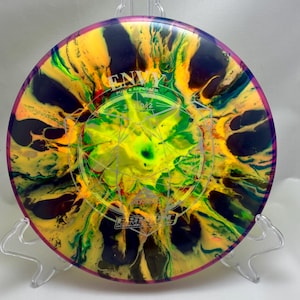 May include: A colorful disc golf disc with a vibrant, swirling pattern of yellow, green, and dark blue. The disc features the word "ENVY" and other text, with a geometric design in the center. The disc is displayed on a clear stand.