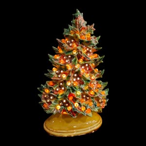 May include: A ceramic Christmas tree with green leaves, red berries, and warm orange lights. The tree sits on a yellow base and is illuminated against a black background. A festive holiday decoration.