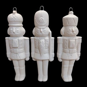 May include: Three white ceramic nutcracker ornaments with silver metal loops for hanging. Each ornament features a different design of a soldier with a mustache, hat, and uniform. The ornaments are set against a black background.