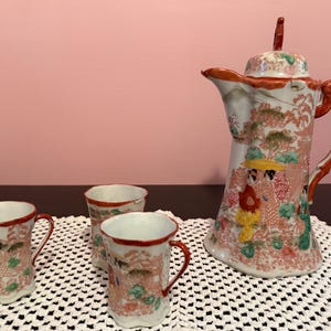 Puede incluir: Un juego de té japonés de porcelana vintage con una tetera alta y adornada y tres tazas a juego. El juego está decorado con una escena de paisaje tradicional en tonos rojos, rosas y verdes, con borde y asa rojos.