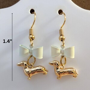 May include: Gold-coloured dachshund dog earrings with white bow accents. The earrings are 3.5 cm in length and feature gold-coloured ear hooks. The earrings are designed for pierced ears, a charming accessory.