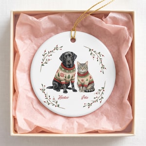 May include: A round white ceramic ornament featuring a black Labrador and a tabby cat, both wearing matching cream, red, and green holiday sweaters with tree patterns. The names Hunter and Pete are printed below them, framed by red berry sprigs.