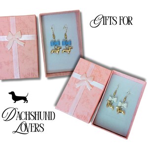 May include: Pink gift boxes with white ribbon and bow, containing dachshund earrings. One pair has blue bows, the other white. The text "Gifts For" and "Dachshund Lovers" is also visible.