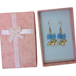 May include: Gold-coloured dachshund earrings with blue bows, displayed in a pink gift box tied with a white ribbon. The earrings are inside a light blue box. The box is closed, ready for gifting.