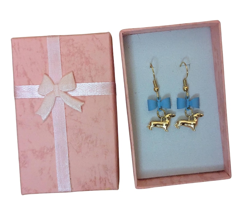 May include: Gold-coloured dachshund earrings with blue bows, displayed in a pink gift box tied with a white ribbon. The earrings are inside a light blue box. The box is closed, ready for gifting.