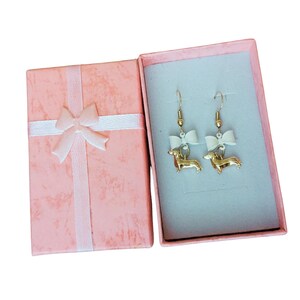 May include: Gold-coloured dachshund dog earrings with white bows, presented in an open pink gift box with a white ribbon and bow. The earrings have gold-coloured hooks.
