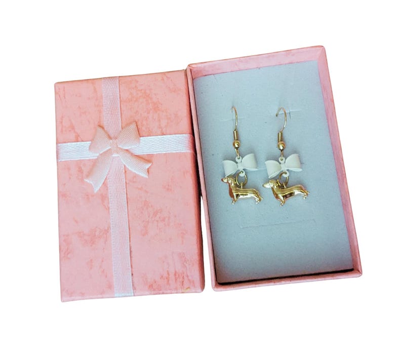 May include: Gold-coloured dachshund dog earrings with white bows, presented in an open pink gift box with a white ribbon and bow. The earrings have gold-coloured hooks.