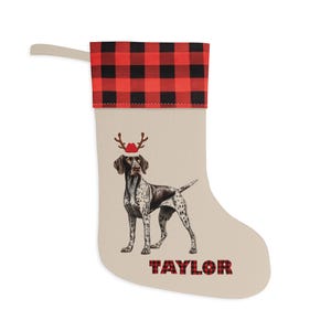 May include: A personalized Christmas stocking with a red and black buffalo plaid cuff. The stocking features a German Shorthaired Pointer dog wearing a Santa hat and reindeer antlers. The name "TAYLOR" is printed on the stocking.