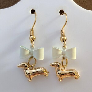 May include: A pair of gold-coloured dachshund dog earrings with a light-coloured bow above each dog. The earrings have gold-coloured hooks and are displayed against a white background.