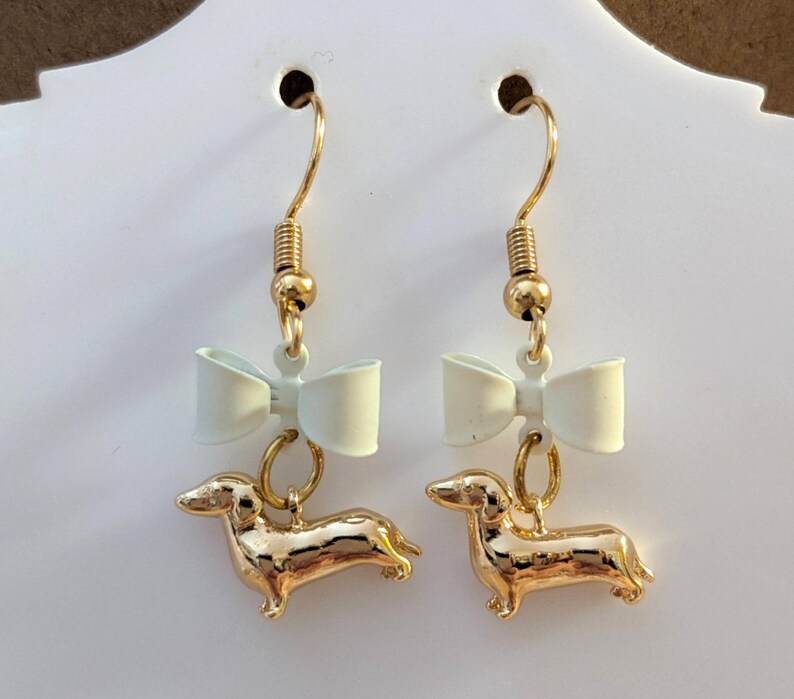 May include: A pair of gold-coloured dachshund dog earrings with a light-coloured bow above each dog. The earrings have gold-coloured hooks and are displayed against a white background.