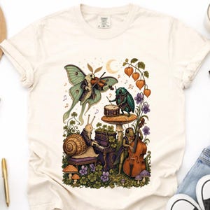 以下が含まれることがあります： クリーム色のTシャツ。バイオリンを弾く蛾、キノコの上でドラムを叩く甲虫、ピアノを弾くカタツムリ、コントラバスを弾くバッタが描かれたグラフィックイラストがプリントされており、周囲には花々と三日月が配置されています。