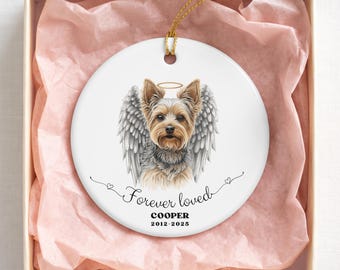 May include: A round white ceramic ornament featuring a detailed illustration of a Yorkshire Terrier with angel wings and a halo. Below the dog, the text reads Forever loved, COOPER, and 2012-2025. The ornament hangs by a gold string.