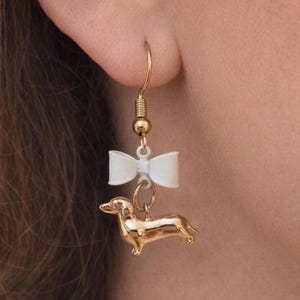 May include: A gold-coloured dachshund dog charm earring with a white bow accent. The earring is suspended from a gold-coloured hook. The earring is shown close up against a blurred background.