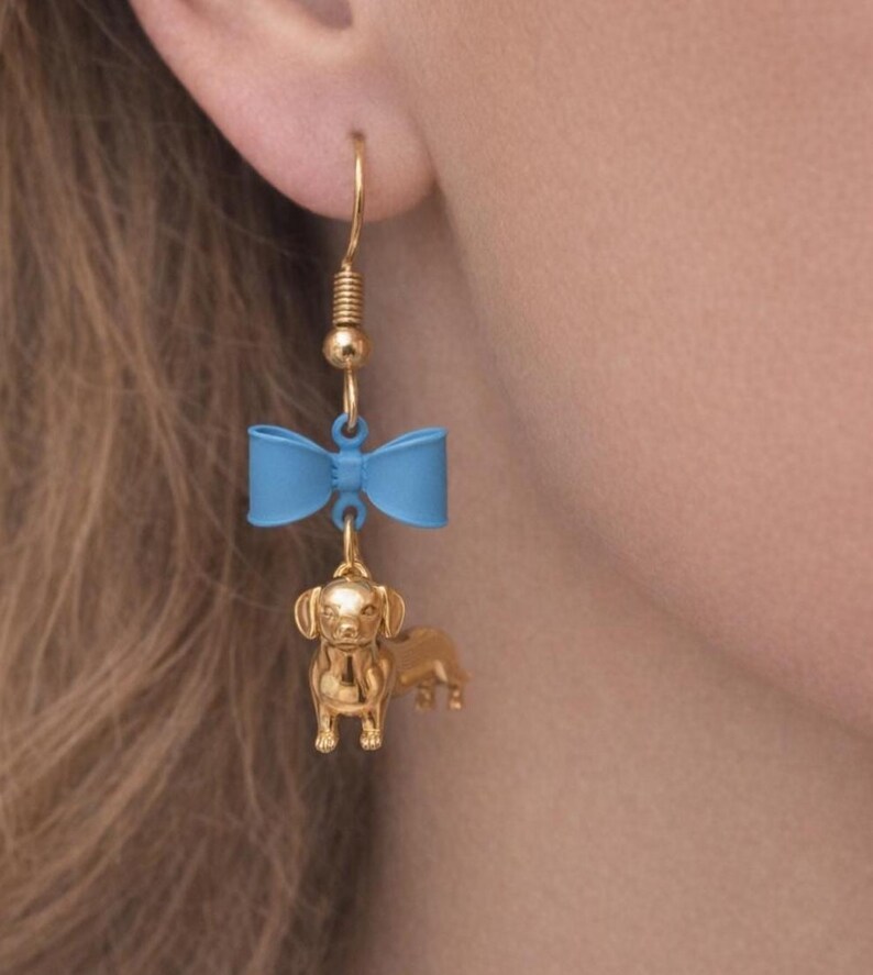 May include: Gold-coloured dachshund dog earrings with a blue bow accent. The earrings have a gold-coloured hook and a detailed miniature dachshund charm. The bow is a light blue colour.