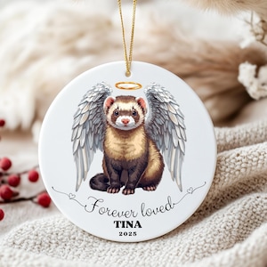 May include: A round white ceramic ornament featuring a brown and tan ferret with large grey angel wings and a golden halo. Below the ferret, the text reads Forever loved, TINA, and 2025 in black script and serif fonts. A gold string is attached.