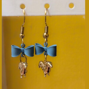 May include: Dangle earrings with a light blue bow and a gold-coloured squirrel charm. The earrings have gold-coloured hooks and a spring detail above the bow. The background is a solid yellow colour.