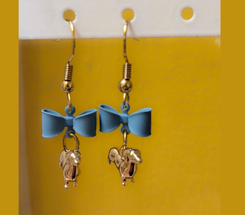 May include: Dangle earrings with a light blue bow and a gold-coloured squirrel charm. The earrings have gold-coloured hooks and a spring detail above the bow. The background is a solid yellow colour.