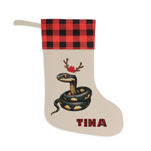 May include: A beige Christmas stocking featuring a red and black buffalo plaid cuff and a hanging loop. The center displays a coiled black and yellow snake wearing small brown reindeer antlers and a red hat. The name TINA is printed in red and black plaid.