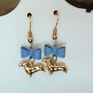 May include: A pair of gold-coloured dachshund dog earrings with blue bows. The earrings have gold-coloured hooks and are suspended from a gold-coloured ball and spring. The dachshunds are in a standing position.