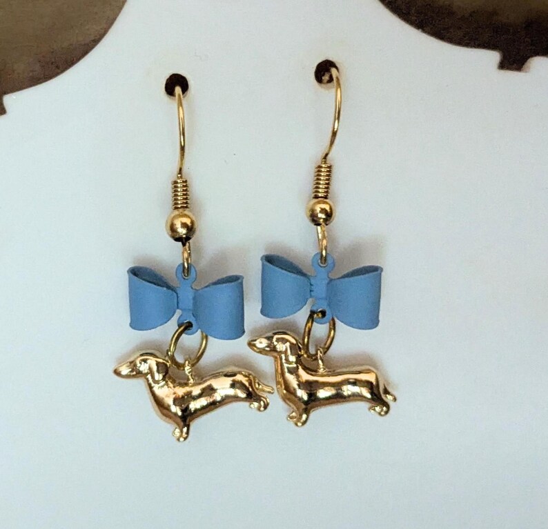 May include: A pair of gold-coloured dachshund dog earrings with blue bows. The earrings have gold-coloured hooks and are suspended from a gold-coloured ball and spring. The dachshunds are in a standing position.