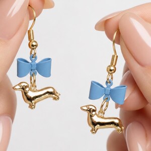May include: Gold-coloured dachshund dog earrings with a light blue bow accent. The earrings have a gold-coloured hook and are dangling. The earrings are held up by a person's hand.